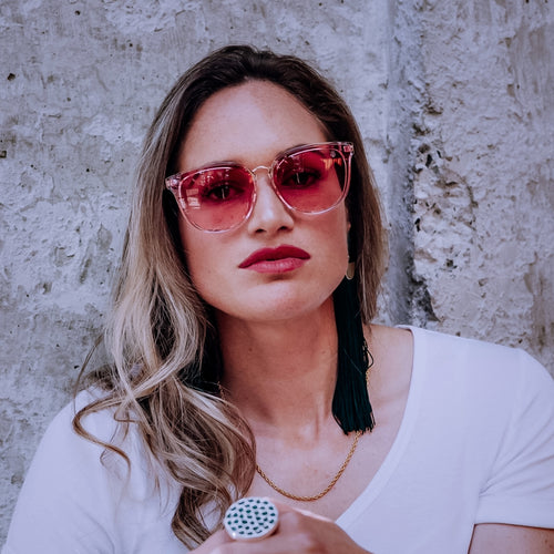 Woman wearing pink Teuila Fatupaito sunglasses Mess Arounds and a white top against a textured wall.